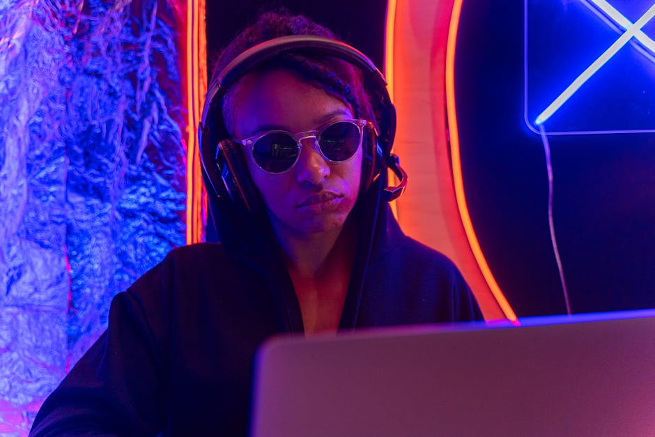 Person in sunglasses and headphones using a laptop in a neon-lit setting.
